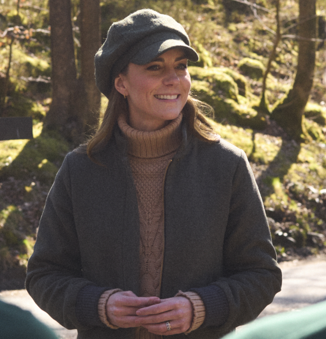 Kate Middleton wearing a tan sweater and green cap smiling in front of some trees