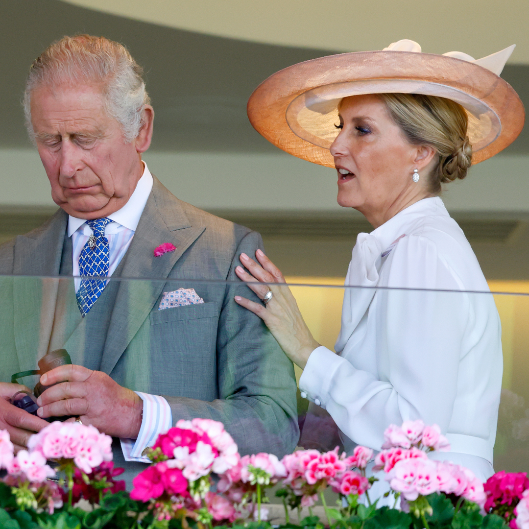 King Charles wears a gray suit and Duchess Sophie, who is wearing a white shirt dress and a wide-brimmed hat, touches his shoudler